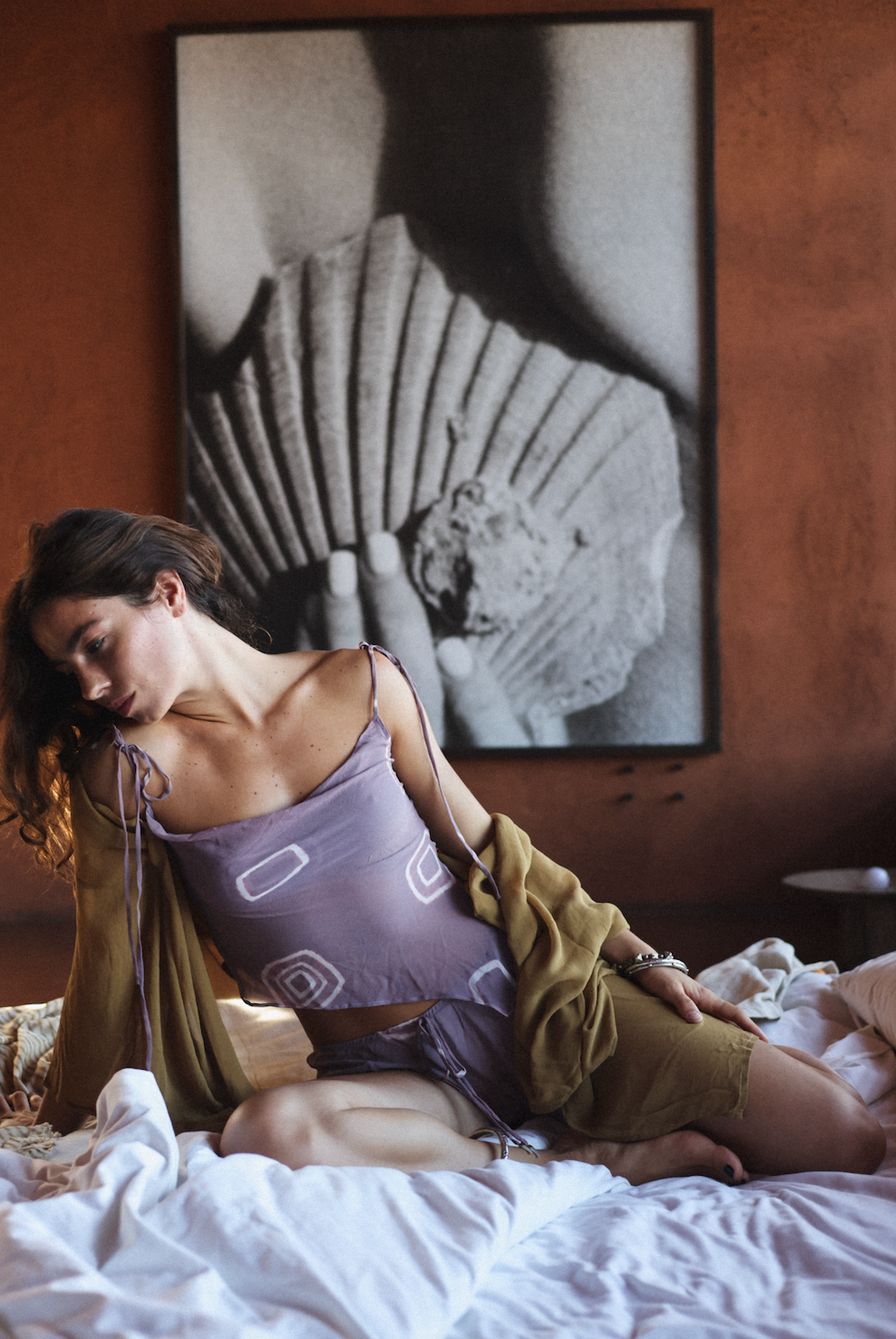 Woman lying on a bed wearing a purple top and green jacket with a large framed black and white abstract painting in the background.